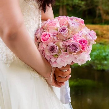 Das Bild zeigt den unteren Oberkörper und die Hände einer Braut, die ein elegantes, weißes Brautkleid trägt. Das Kleid ist aus feinem Stoff mit Spitzenapplikationen im Oberteil und einem fließenden Rockteil. Die Braut hält ihren Brautstrauß mit beiden Händen auf dem Rücken. Der Strauß besteht aus verschiedenen Rosatönen, darunter hellrosa, kräftiges Pink und zartes Lavendel. Die Blumen sind eng gebunden, vermutlich Rosen und Hortensien, und mit Perlen verziert. Am Handgelenk der Braut ist ein feines Armband zu sehen, an der linken Hand trägt sie einen Ehering. Der Hintergrund des Bildes ist entfernt bzw. transparent.
