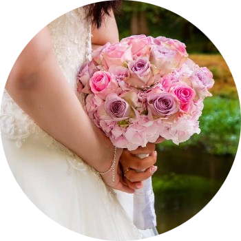 Das Bild zeigt den unteren Oberkörper und die Hände einer Braut, die ein elegantes, weißes Brautkleid trägt. Das Kleid ist aus feinem Stoff mit Spitzenapplikationen im Oberteil und einem fließenden Rockteil. Die Braut hält ihren Brautstrauß mit beiden Händen auf dem Rücken. Der Strauß besteht aus verschiedenen Rosatönen, darunter hellrosa, kräftiges Pink und zartes Lavendel. Die Blumen sind eng gebunden, vermutlich Rosen und Hortensien, und mit Perlen verziert. Am Handgelenk der Braut ist ein feines Armband zu sehen, an der linken Hand trägt sie einen Ehering. Der Hintergrund des Bildes ist entfernt bzw. transparent.