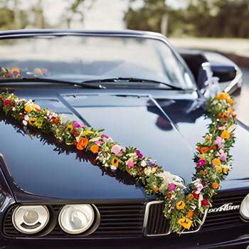 Das Bild zeigt die Front eines dunkelblauen Oldtimers, vermutlich ein Hochzeitsauto. Über die Motorhaube ist ein großes, V-förmiges Blumengesteck gespannt. Die Blumengirlande besteht aus vielen bunten, frischen Blumen – darunter Gelb, Orange, Rosa, Weiß, Lila und Rot – sowie viel grünem Laub. Die Blumen wirken wie eine Mischung aus Garten- und Wiesenblumen, was dem Arrangement einen natürlichen und festlichen Charakter verleiht. Die Spiegelung auf dem Lack zeigt Gebäude und Himmel, was auf sonniges Wetter hinweist. Im Hintergrund ist leicht unscharf die Straße zu erkennen. Das Auto vermittelt durch die Dekoration deutlich, dass es für eine Hochzeit vorbereitet wurde.