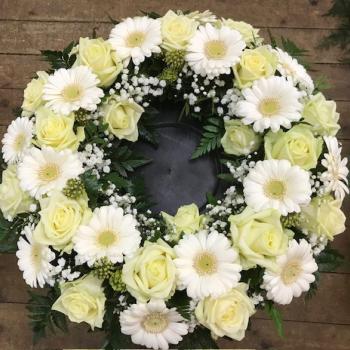 Auf dem Bild ist ein runder, kunstvoll arrangierter Blumenkranz zu sehen. Der Kranz besteht hauptsächlich aus cremefarbenen Rosen und weißen Gerbera, die regelmäßig im Kreis angeordnet sind. Zwischen den großen Blüten befinden sich kleinere weiße Blüten, wahrscheinlich Schleierkraut, sowie grünes Blattwerk. Der Kranz liegt auf einem hölzernen Untergrund und hat in der Mitte eine offene Fläche, in der eine dunkle Kunststoff- oder Gummiunterlage sichtbar ist. Die Farben des Kranzes wirken zart und harmonisch – Weiß, Creme und Grün dominieren. Die Anordnung und die Farbgebung lassen darauf schließen, dass es sich um einen Trauerkranz handelt.