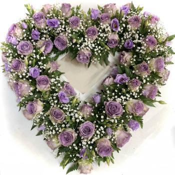   Ein herzförmiges Blumenarrangement liegt auf einem weißen Untergrund. Das Herz besteht aus einer dichten Umrandung aus frischen, lilafarbenen Rosen, zarten violetten Lisianthusblüten und kleinen weißen Schleierkrautblüten. Die Blumen sind kunstvoll miteinander verflochten und von dunkelgrünem Blattwerk umrahmt. Die Mitte des Herzens ist offen, wodurch die Form besonders betont wird. Die Farben wirken harmonisch und sanft, was dem Arrangement eine würdevolle, ruhige und liebevolle Ausstrahlung verleiht – passend für einen feierlichen oder erinnernden Anlass wie eine Trauerfeier oder Gedenkveranstaltung.