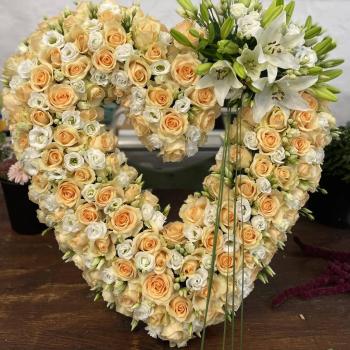 Das Bild zeigt ein großes, herzförmiges Blumenarrangement, das auf einer Holzoberfläche steht. Die Herzform besteht hauptsächlich aus dicht gesteckten Rosen in zarten Apricot- und Cremefarben sowie kleinen weißen Blüten, vermutlich Lisianthus. Rechts oben am Herz ist ein auffälliger Akzent aus weißen Lilien gesetzt, die von grünen Knospen und länglichen Blättern umgeben sind. Einige lange, gerade grüne Grashalme hängen dekorativ nach unten. Im Hintergrund ist eine weiße Wand zu sehen sowie teilweise andere Blumengefäße. Die Komposition wirkt elegant und sorgfältig gestaltet, vermutlich für einen feierlichen Anlass wie eine Trauerfeier oder Hochzeit.